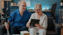 Elder Couple Looking At Tablet Screen In Living Room, Enjoying Free Time Together. Retired Man With Crutches And Woman With Digital Device Browsing Internet To Have Leisure Activity. Modern People