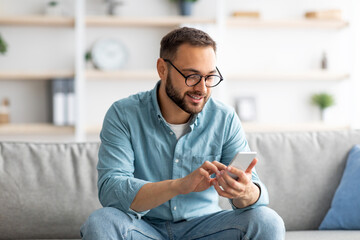 Poster - Positive young guy in eyeglasses sitting on sofa with mobile phone and texting to his friend, indoors