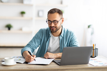 Poster - Young Caucasian businessman taking notes while working on laptop computer at home office
