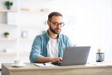 Fototapete - Young Caucasian man in eyeglasses working online, sitting at desk and using laptop from home office