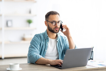 Poster - Portrait of happy young guy using laptop, speaking on cellphone, having freelance job at home