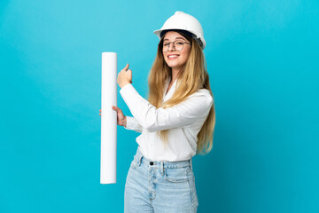 Young architect woman with helmet and holding blueprints isolated on blue background pointing back