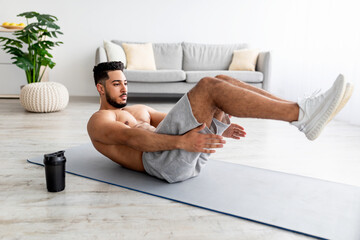 Wall Mural - Shirtless young Arab man working out core muscles, doing abs exercises on yoga mat at home, full length