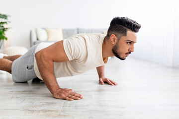Wall Mural - Millennial Arab man doing push ups, standing in plank pose, doing strength exercises at home. Domestic workout concept