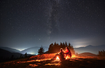 Wall Mural - Side view of pair sitting one behind other on meadow near bright flame of bonfire and watching surrounding beauty of evening starry sky. Mountain hills and lighted hamlets in the distance.