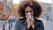 Sick ill african american woman with curly hair blowing runny nose into tissue paper outdoors cold allergies respiratory virus girl suffers from contagious rhinitis symptom autumn in city flu pandemic