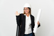 © luismolinero - Middle age architect woman with helmet and holding blueprints over isolated background saluting with hand with happy expression
