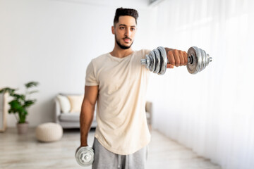 Wall Mural - Portrait of handsome young Arab guy exercising with dumbbells at home during covid-19 lockdown, selective focus