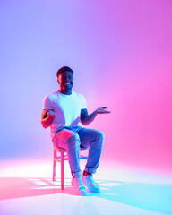 Poster - Joyful African American man sitting on chair, presenting empty space for your ad, smiling at camera in neon light