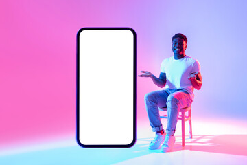 Poster - Positive black man sitting on chair near huge cellphone with empty white screen in neon light, mockup for mobile app