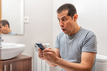Latin Man Receiving Surprising News On Line In A Smart Phone At The Bathroom.