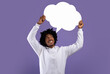 Handsome black teenager holding empty speech bubble above his head on violet studio background, mockup