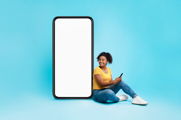 Wall Mural - Cheerful black woman sitting near huge smartphone with empty screen, holding mobile device on blue background, mockup