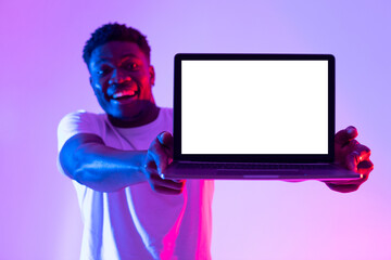 Poster - Cheerful black guy holding laptop pc with empty white screen with space for mockup, promoting new website in neon light