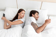 Upset european wife jealous and looks at phone of smiling husband on white bed in bedroom interior