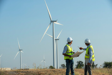 Back view of two engineers discussing against turbines on wind farm while walking along road on sunny day