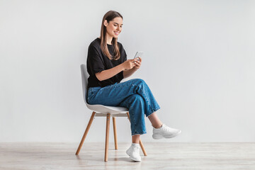 Poster - Happy young lady sitting on chair with smartphone, texting to friend, sending message, browsing web against white wall