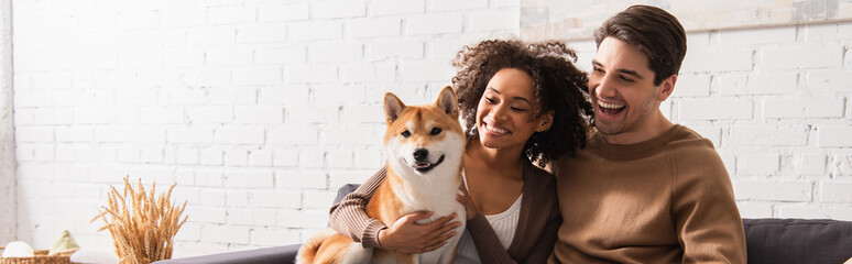 Wall Mural - Happy man looking at smiling african american girlfriend with shiba inu at home, banner