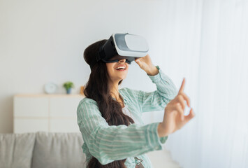 Wall Mural - Young Indian woman in VR glasses exploring cyberspace, touching imaginary screen, playing video game at home