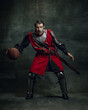 Portrait of medieval warrior or knight with dirty wounded face playing with basketball ball isolated over dark background. Comparison of eras, history