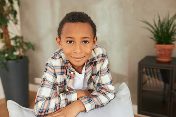 Close-up of handsome little man leaning on back of sofa, looking at camera smiling, having cute face, brown short curly hair and adorable charismatic smile, standing against room interior