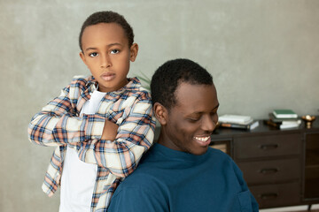 Wall Mural - Indoor picture of two siblings, little serious elementary school boy with hands crossed leaning against his 20-years-old brother sitting in chair against living-room interior, smiling looking down