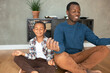 © Anatoliy Karlyuk - Portrait of father and son of black ethnicity fooling around pretending meditating, sitting in lotus posture with eyes closed, smiling, having fun, both dressed casually in brown jeans