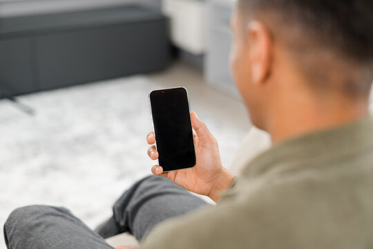 Back view of multiracial man sitting on sofa in office, using mobile phone for checking social nets, typing message for family, girlfriend, business partner, views photos, watches videos