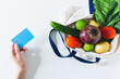 Female hand holding a credit card and eco bag with vegetables on white background. Flat lay, top view, copy space.