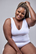 © Roman - Plump, plus size black woman in white bodysuit laughing, sitting in studio over white background, laughing. Concept of body positive, equality, people diversity. copy space, free space. portrait
