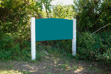 Blank Green Wooden Sign On White Posts With Large Bushes In The Background