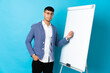 © luismolinero - Young Colombian man isolated on blue background giving a presentation on white board and pointing it