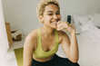 © shurkin_son - Female of 20s with dark skin isolated against blurred background of bedroom, unmade bad and interior details, posing with happy toothy smile, dressed in sportswear, looking stylish with blonde curls