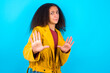 Afraid African teenager girl wearing yellow jacket over blue background, makes terrified expression and stop gesture with both hands saying: Stay there. Panic concept.
