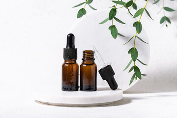 Two glass bottles with skin care serum on white marble podium with fresh green eucalyptus leaves close up. Natural herbal cosmetic banner.