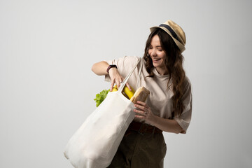 Wall Mural - Smiling young woman in beige t-shirt and a hst with a mesh eco bag full of vegetables and on a white studio background. Sustainable lifestyle. Eco friendly concept. Zero waste.