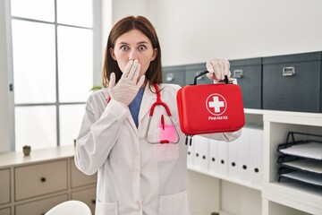 Canvas Print - Young doctor woman holding first aid kit covering mouth with hand, shocked and afraid for mistake. surprised expression