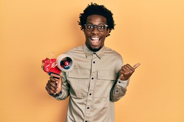 Poster - Young african american man holding packing tape machine pointing thumb up to the side smiling happy with open mouth