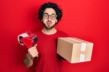 Poster - Handsome hispanic man holding packing tape and cardboard box puffing cheeks with funny face. mouth inflated with air, catching air.