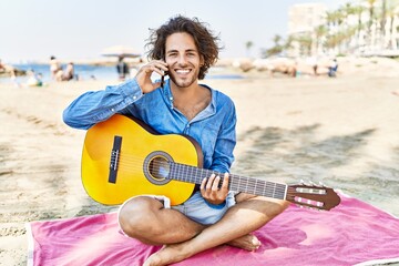 Canvas Print - Young hispanic man playing guitar and talking on the smartphone sitting on sand at the beach.