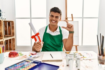 Poster - Young hispanic man at art studio holding degree smiling and confident gesturing with hand doing small size sign with fingers looking and the camera. measure concept.