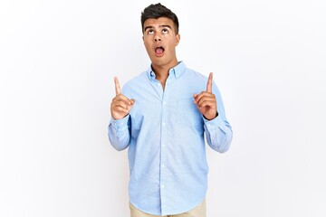 Sticker - Young hispanic man wearing business shirt standing over isolated background amazed and surprised looking up and pointing with fingers and raised arms.