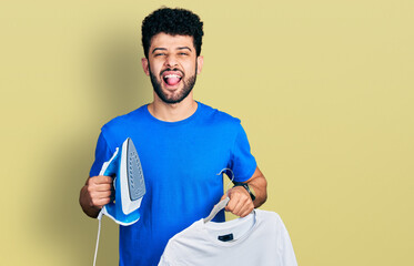 Canvas Print - Young arab man with beard holding electric steam iron and white t shirt sticking tongue out happy with funny expression.