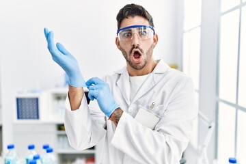 Sticker - Young hispanic man working at scientist laboratory putting gloves on afraid and shocked with surprise and amazed expression, fear and excited face.