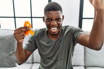 Canvas Print - Young african american man holding awareness orange ribbon sitting on the sofa annoyed and frustrated shouting with anger, yelling crazy with anger and hand raised