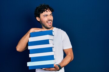 Wall Mural - Young hispanic man holding a pile of books angry and mad screaming frustrated and furious, shouting with anger. rage and aggressive concept.