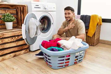 Wall Mural - Young handsome man putting dirty laundry into washing machine with a big smile on face, pointing with hand and finger to the side looking at the camera.