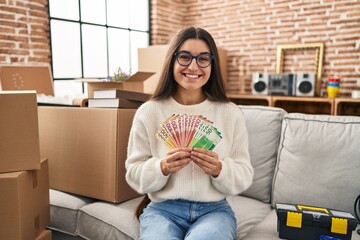 Poster - Young hispanic woman sitting on the sofa at new home holding money smiling with a happy and cool smile on face. showing teeth.