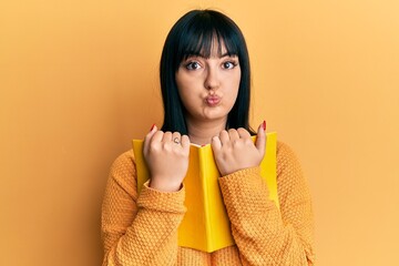 Poster - Young hispanic woman holding book puffing cheeks with funny face. mouth inflated with air, catching air.