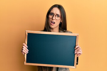 Poster - Young hispanic girl holding blackboard in shock face, looking skeptical and sarcastic, surprised with open mouth